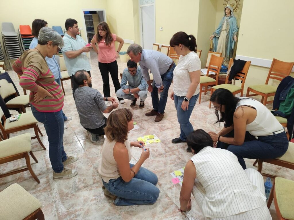 Participantes del curso agachados en círculo en el salón parroquial trabajando con notas adhesivas en el suelo.