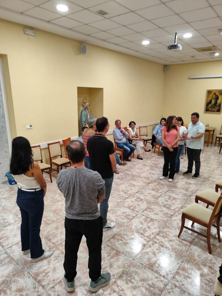 Varios adultos de pie y sentados en círculo conversando en una sala parroquial.