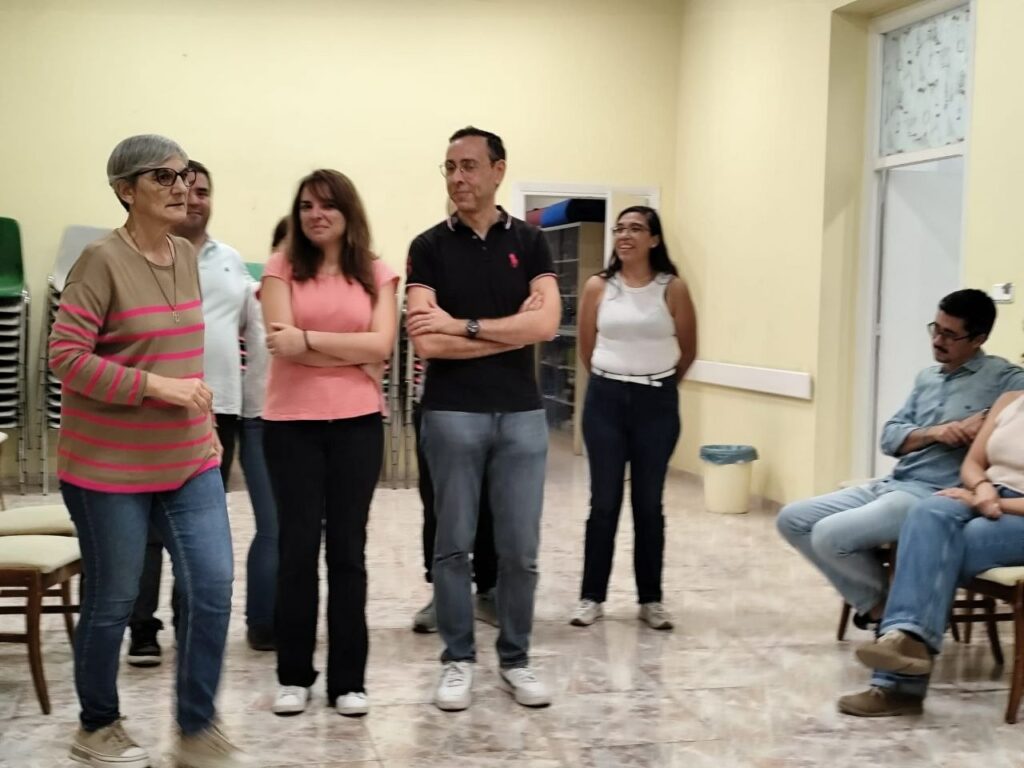 Grupo de hombres y mujeres de pie sonriendo y conversando en una sala con sillas al fondo.