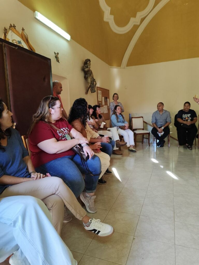 Grupo de adultos sentados en sillas conversando en una sala parroquial iluminada.