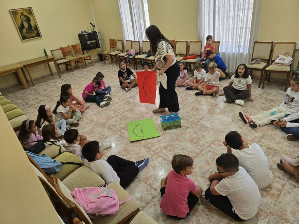 Grupo de niños sentados en el suelo alrededor de una catequista con cartulinas.