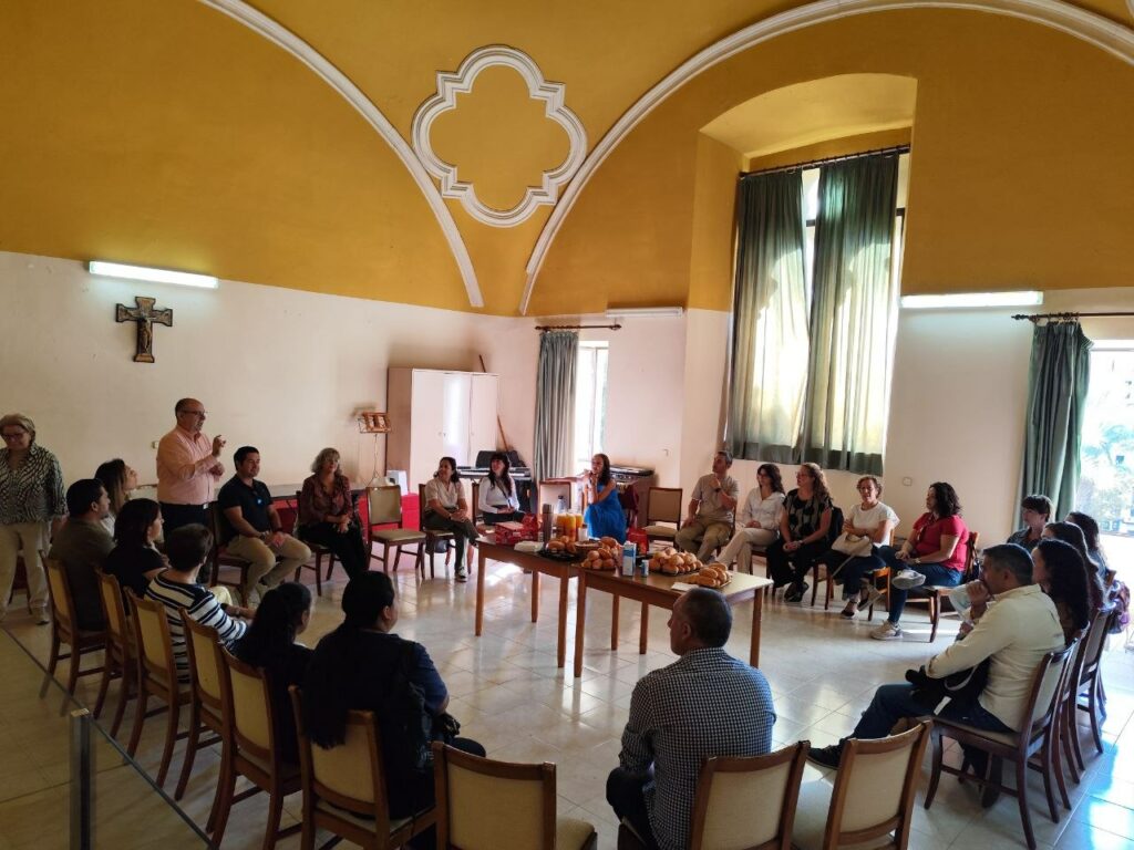 Grupo amplio de adultos en círculo alrededor de mesas con comida y bebida en una sala.